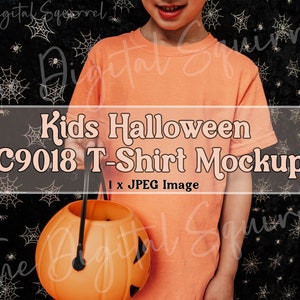 May include: A child wearing an orange t-shirt holds a pumpkin-shaped trick-or-treat bucket. The background is black with white spiderwebs.