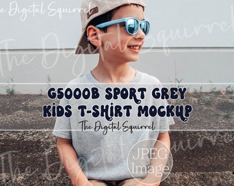May include: A child wearing a light gray t-shirt, blue sunglasses, and a beige baseball cap. The shirt has the text "G5000B SPORT GREY KIDS T-SHIRT MOCKUP" in dark blue. The image also includes the text "The Digital Squirrel" and "JPEG Image."