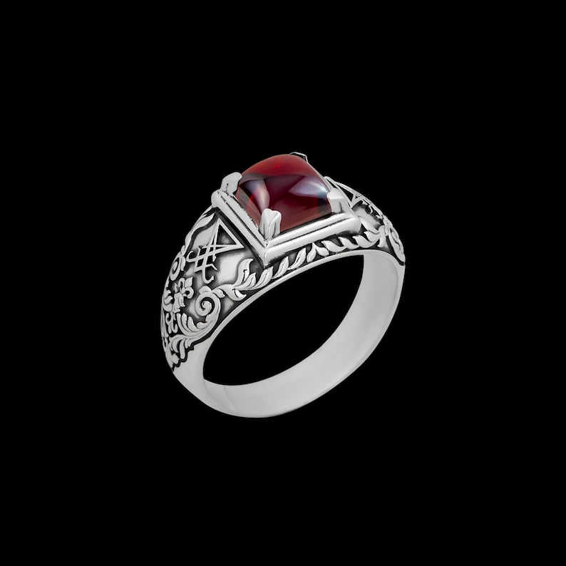 May include: A silver ring with an ornate design and a red gemstone. The ring features a square-shaped stone set in a silver bezel.