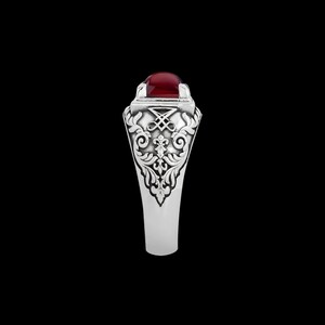 May include: A silver ring with an ornate design and a red gemstone. The ring has a detailed floral pattern and a square-shaped setting for the gemstone.