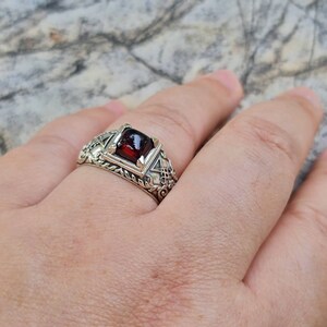 May include: A silver ring with a red gemstone set in a square bezel. The ring has intricate details and a rope-like band.