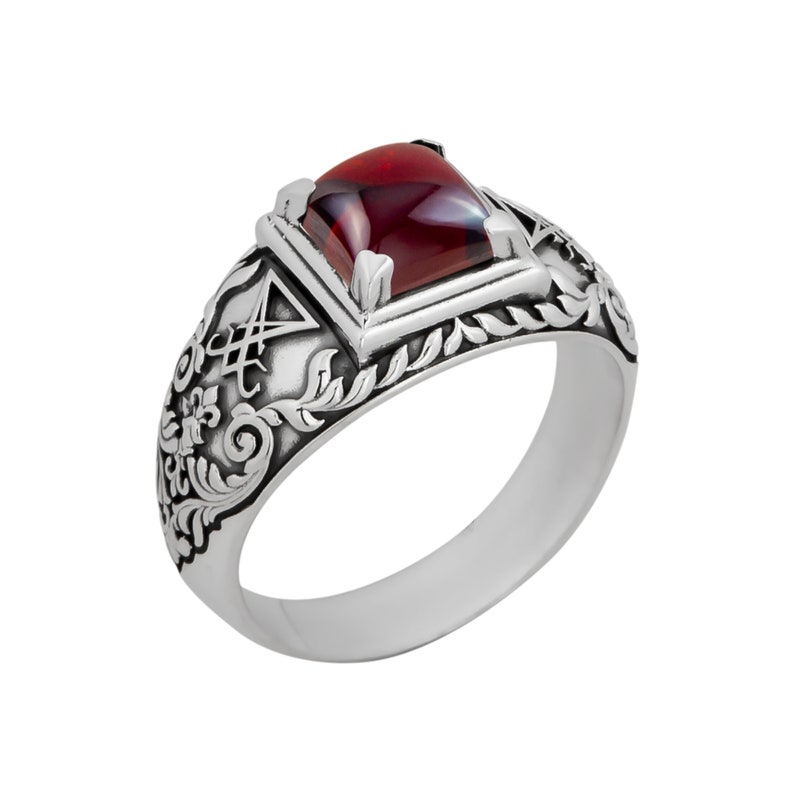 May include: A silver ring with a red gemstone set in a square bezel. The ring features an intricate design of leaves and swirls.