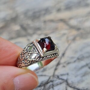 May include: A silver ring with an ornate design featuring a dark red gemstone. The ring has a detailed floral pattern and a symbol resembling a pentagram.