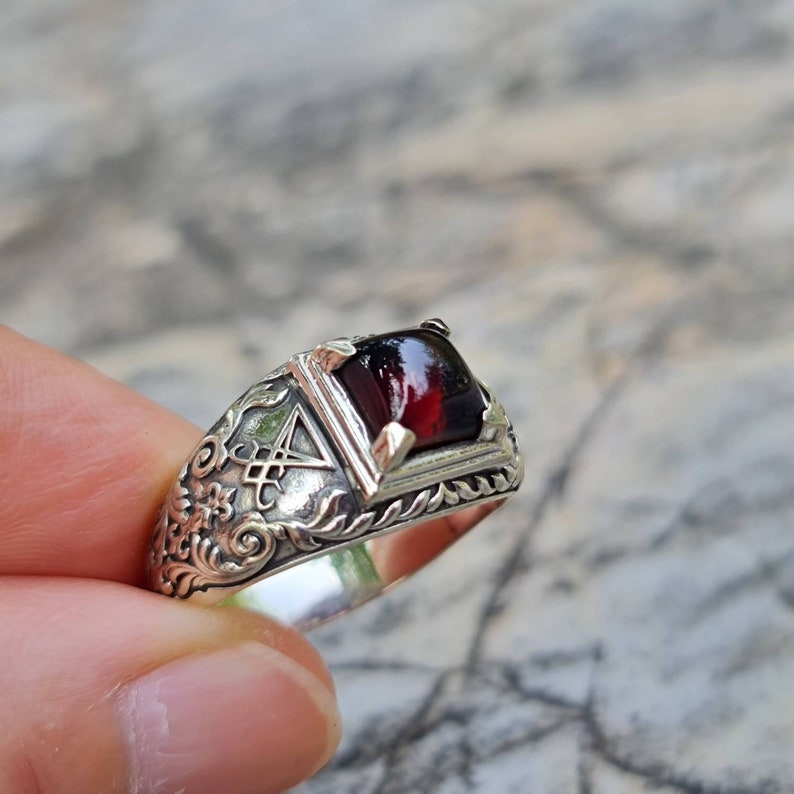May include: A silver ring with an ornate design featuring a dark red gemstone. The ring has a detailed floral pattern and a symbol resembling a pentagram.