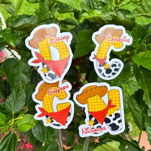 May include: Four decorative garden stakes featuring a cartoon design. Each stake has a letter with a cowboy hat, red bandana, and a name: Danny, Catherine, Greyson, and Nicholas. The letters are yellow with a grid pattern.