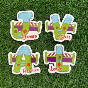 May include: Four colorful name stickers with a cartoon design. Each sticker features a letter in a unique font, with the names "James", "Violet", "Alexander", and "Logan" written below. The stickers are on a green background.