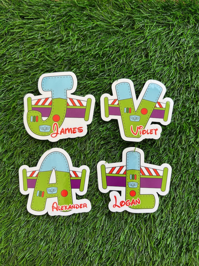 May include: Four colorful name stickers with a cartoon design. Each sticker features a letter in a unique font, with the names "James", "Violet", "Alexander", and "Logan" written below. The stickers are on a green background.
