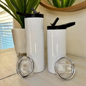 Puede incluir: Dos vasos aislados blancos con tapas negras y tapas de pl&aacute;stico transparente. Los vasos son cil&iacute;ndricos y tienen un acabado brillante. Una tapa est&aacute; abierta, revelando la boquilla para beber. Los vasos se exhiben sobre una superficie de madera clara.