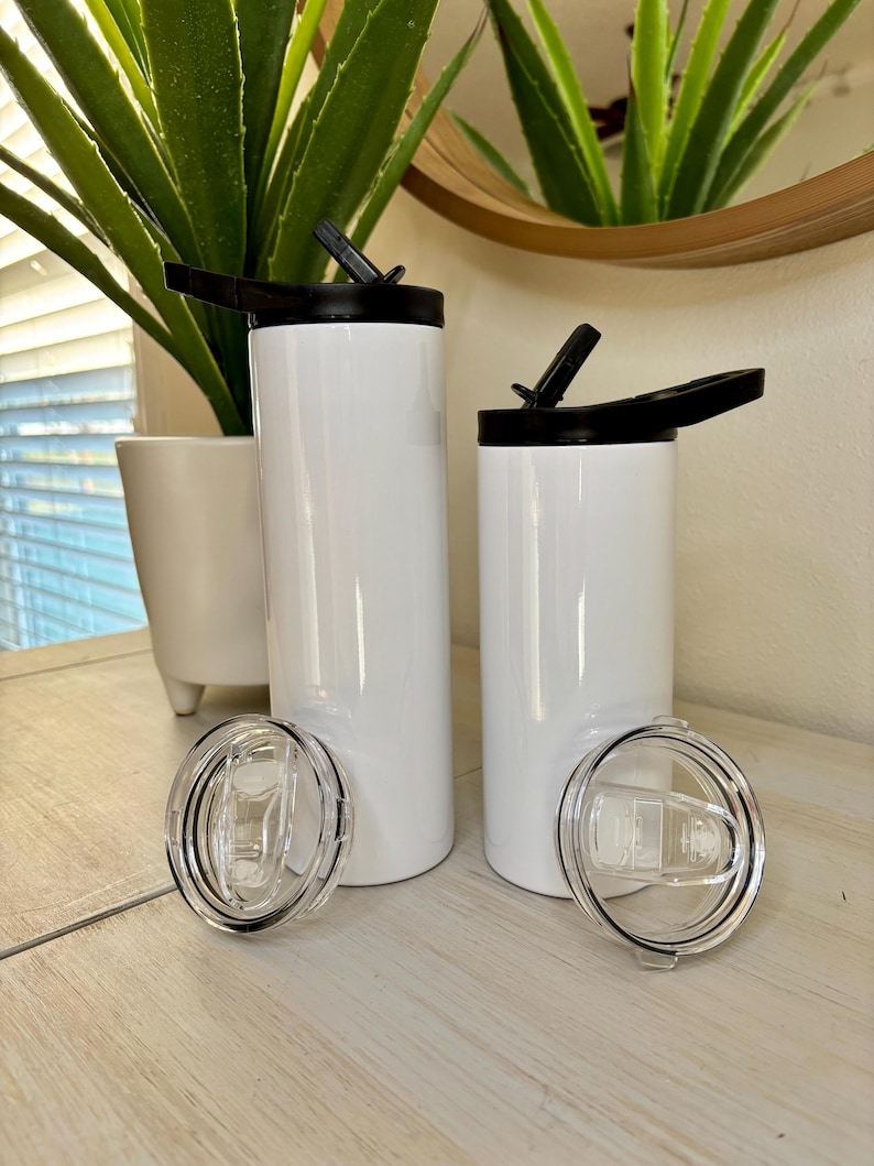 Puede incluir: Dos vasos aislados blancos con tapas negras y tapas de pl&aacute;stico transparente. Los vasos son cil&iacute;ndricos y tienen un acabado brillante. Una tapa est&aacute; abierta, revelando la boquilla para beber. Los vasos se exhiben sobre una superficie de madera clara.