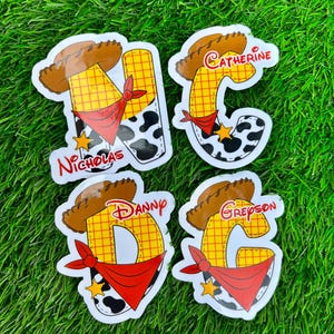 May include: Four custom stickers with a cowboy theme. Each sticker features a letter, a cowboy hat, a red bandana, and a cow-print design. The names "Nicholas", "Catherine", "Danny", and "Greyson" are printed on the stickers.