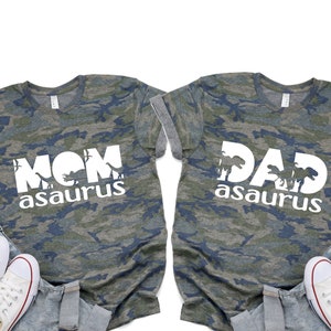 May include: Two camouflage t-shirts with the text "MOM asaurus" and "DAD asaurus" in white. The shirts are laid out on a light blue denim background with white sneakers.