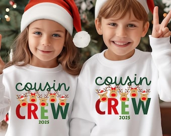 May include: Two children wearing white sweatshirts and red Santa hats pose in front of a Christmas tree. The sweatshirts read "Cousin Crew 2025" in green and red lettering, with reindeer graphics.