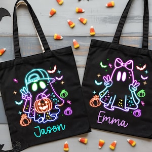 May include: Two black Halloween tote bags with neon ghost designs, pumpkins, and the names "Jason" and "Emma." The bags are surrounded by candy corn, paper bats, and small pumpkins, creating a festive scene.