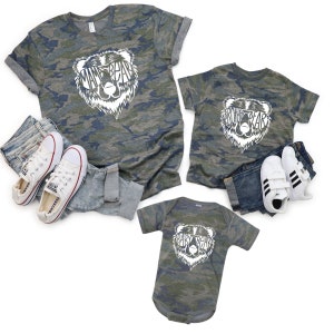 May include: Camouflage-patterned t-shirts and a baby bodysuit with bear graphics. The shirts say "Mama Bear" and "Brother Bear", while the bodysuit says "Baby Bear". White sneakers and jeans are also in the image.
