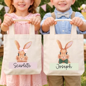 May include: Two canvas tote bags with bunny illustrations. One bag features a pink bow and the name "Scarlette" on a pink and white checkered banner. The other bag has a green bow tie and the name "Joseph" on a green and white checkered banner.