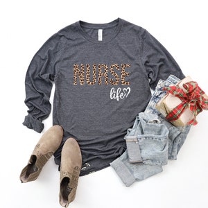 K&ouml;nnte beinhalten: Ein dunkelgraues Langarmshirt mit dem Aufdruck "NURSE life" auf der Vorderseite. Das Wort "NURSE" ist in einem Leopardenmuster gehalten. Das Shirt wird mit hellblauen Jeans und braunen Stiefeletten kombiniert.