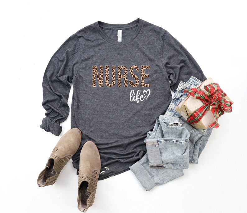 K&ouml;nnte beinhalten: Ein dunkelgraues Langarmshirt mit dem Aufdruck "NURSE life" auf der Vorderseite. Das Wort "NURSE" ist in einem Leopardenmuster gehalten. Das Shirt wird mit hellblauen Jeans und braunen Stiefeletten kombiniert.