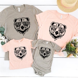 May include: Four t-shirts and a onesie in various colors, each featuring a black bear face design. The bear's face is filled with the words "PAPA BEAR", "MAMA BEAR", "SISTER BEAR", and "BABY BEAR". A straw hat and greenery are in the background.