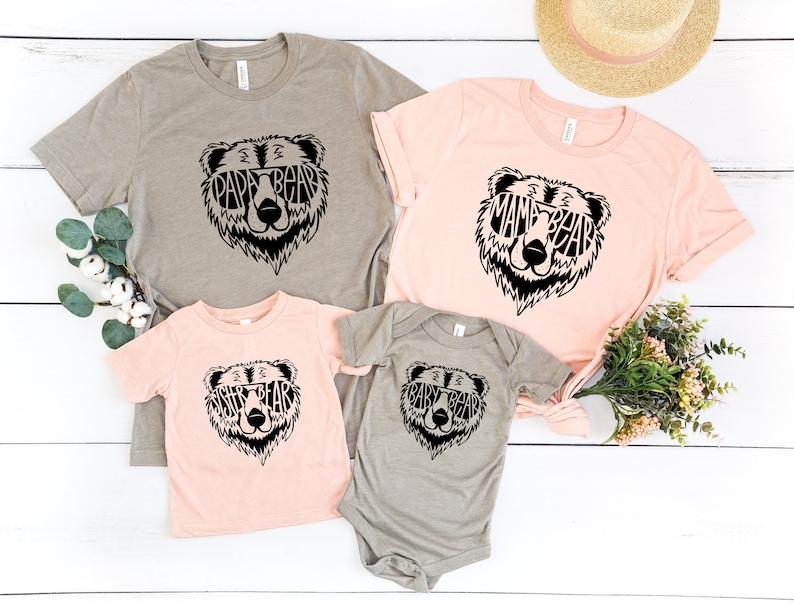 May include: Four t-shirts and a onesie in various colors, each featuring a black bear face design. The bear's face is filled with the words "PAPA BEAR", "MAMA BEAR", "SISTER BEAR", and "BABY BEAR". A straw hat and greenery are in the background.