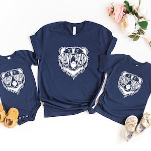May include: Navy blue t-shirts and a onesie with a white bear graphic that says "Mama Bear", "Papa Bear", and "Baby Bear". The shirts are arranged with baby shoes and flowers.