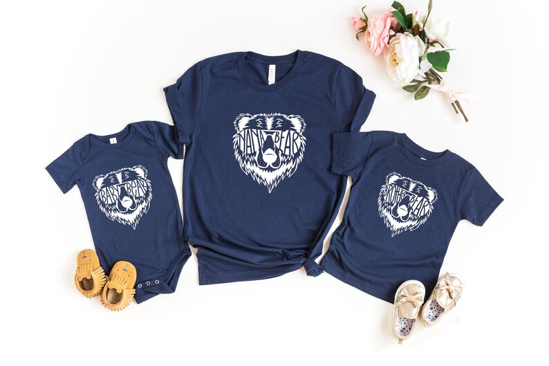 May include: Navy blue t-shirts and a onesie with a white bear graphic that says "Mama Bear", "Papa Bear", and "Baby Bear". The shirts are arranged with baby shoes and flowers.