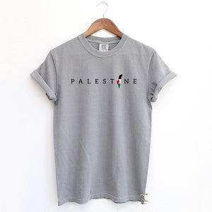 May include: A gray t-shirt with the word "PALESTINE" printed in black letters. The word is centered on the shirt and is accompanied by an outline of the Palestinian territories in black with the Palestinian flag colors inside.