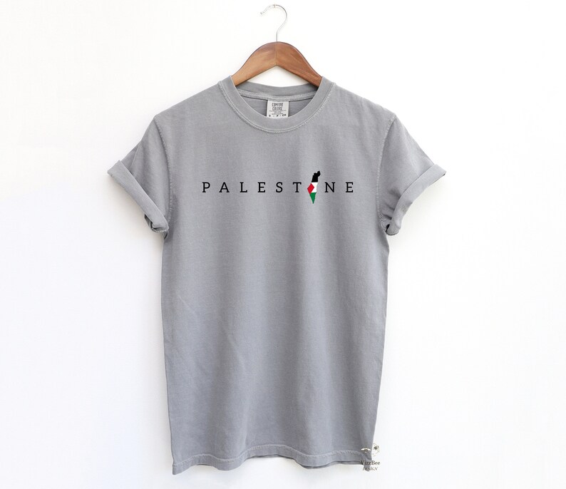 May include: A gray t-shirt with the word "PALESTINE" printed in black letters. The word is centered on the shirt and is accompanied by an outline of the Palestinian territories in black with the Palestinian flag colors inside.