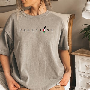May include: A gray t-shirt with the word "PALESTINE" printed in black letters and an outline of the Palestinian territory with the Palestinian flag colors in the center.