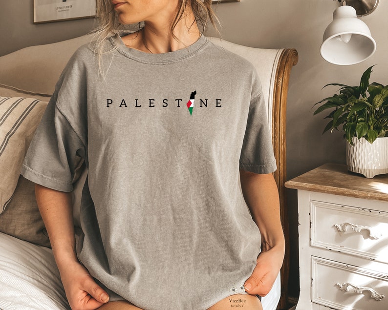 May include: A gray t-shirt with the word "PALESTINE" printed in black letters and an outline of the Palestinian territory with the Palestinian flag colors in the center.