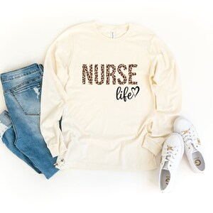 K&ouml;nnte beinhalten: Ein lang&auml;rmeliges, cremefarbenes T-Shirt mit dem Text "NURSE life" in braunem und schwarzem Leopardenmuster. Das Shirt wird mit einer blauen Jeans und wei&szlig;en Turnschuhen kombiniert.