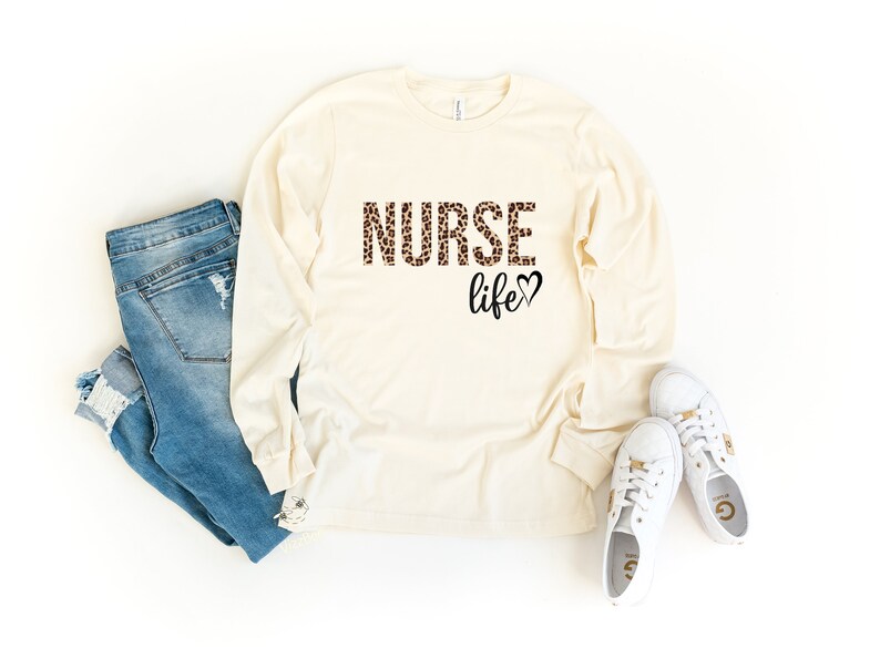 K&ouml;nnte beinhalten: Ein lang&auml;rmeliges, cremefarbenes T-Shirt mit dem Text "NURSE life" in braunem und schwarzem Leopardenmuster. Das Shirt wird mit einer blauen Jeans und wei&szlig;en Turnschuhen kombiniert.