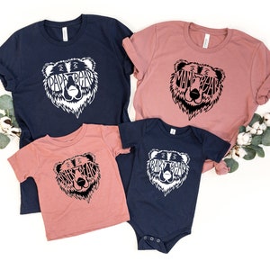 May include: A collection of matching family t-shirts and a onesie. The shirts are navy blue and dusty rose, with a white bear graphic that says "Papa Bear," "Mama Bear," "Sister Bear," and "Baby Bear." The onesie is navy blue.