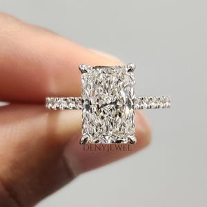 May include: A radiant-cut diamond engagement ring. The ring features a large, rectangular-shaped diamond as the centerpiece, with smaller diamonds set along the band. The ring is held up against a neutral background.