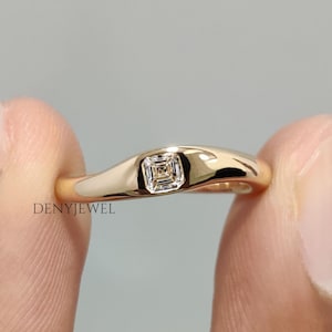 May include: A gold ring featuring a square-cut diamond. The ring's band is smooth and curved, and it is held between two fingers. The ring is a piece of jewellery.