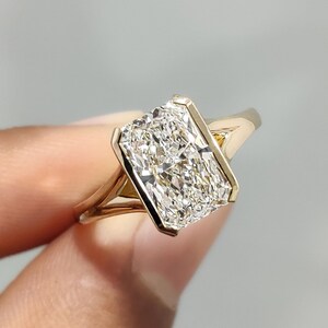 May include: A gold ring featuring a radiant-cut diamond. The rectangular diamond is set within a bezel, highlighting its sparkle. The gold band has a split shank design. The ring is a beautiful piece of jewellery.
