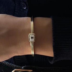 Puede incluir: Un brazalete dorado con un diseño central de forma ovalada. El brazalete presenta una pequeña piedra transparente en el centro del óvalo. El brazalete se lleva en la muñeca, con una rebeca azul oscuro y vaqueros en el fondo.