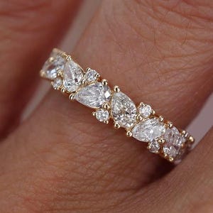 May include: A gold band ring set with multiple clear, sparkling gemstones. The ring features a repeating pattern of marquise and round-cut stones, creating a continuous design. The ring is worn on a finger, showcasing its intricate details and elegant style.