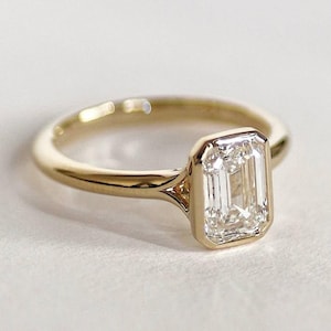 Op de afbeelding: Een gouden ring met een enkele, smaragdgeslepen heldere edelsteen in een gladde zetting. De ringband is van gepolijst geelgoud met een taps toelopend ontwerp dat aansluit op de centrale steenzetting, wat zorgt voor een strakke uitstraling.