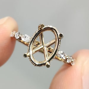 May include: A gold ring with an oval setting, designed to hold a gemstone. The band features small, clear accent stones. The ring is held between two fingers against a neutral background.