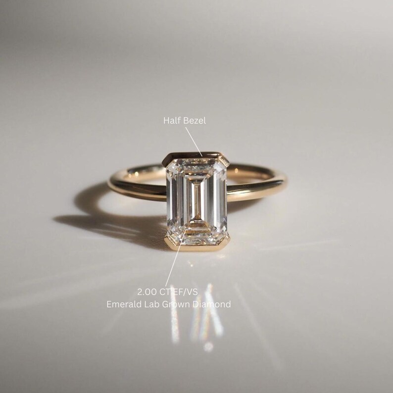 Puede incluir: Un anillo de oro con un diamante de laboratorio de talla esmeralda de 2,00 quilates engastado en una media engaste. El diamante es claro y tiene un brillo brillante. El anillo es simple y elegante.