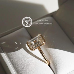 Puede incluir: Un anillo de oro con un gran diamante de laboratorio de talla esmeralda, transparente. El anillo est&aacute; en una caja blanca con el texto "IGI Certified Lab Grown Diamond".