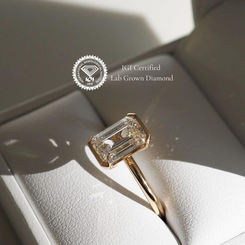 Puede incluir: Un anillo de oro con un gran diamante de laboratorio de talla esmeralda, transparente. El anillo est&aacute; en una caja blanca con el texto "IGI Certified Lab Grown Diamond".