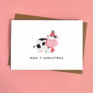 Peut inclure: Carte de vœux blanche avec une vache de dessin animé portant un chapeau et une écharpe rouges, avec le texte "MOO-Y CHRISTMAS". La carte est sur une enveloppe marron, sur fond rose.