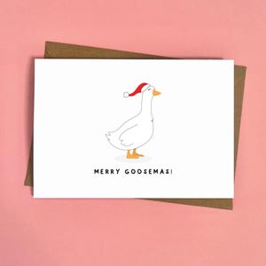 May include: A white greeting card with a cartoon goose wearing a Santa hat. The card has the text "MERRY GOOSEMAS!" in black font. The card is on a brown envelope, set against a pink background.