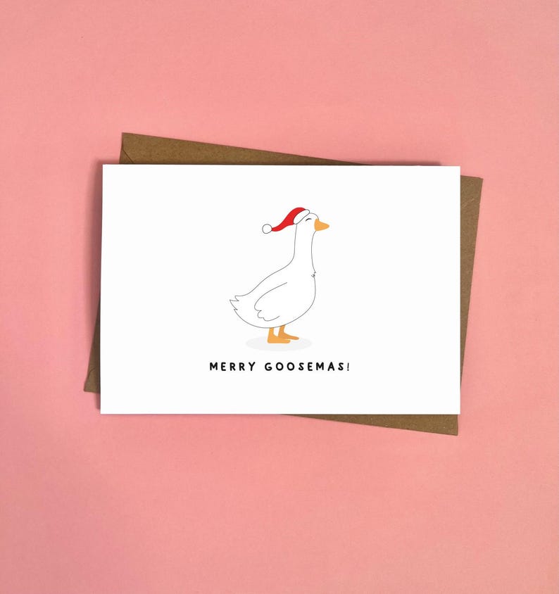 May include: A white greeting card with a cartoon goose wearing a Santa hat. The card has the text "MERRY GOOSEMAS!" in black font. The card is on a brown envelope, set against a pink background.