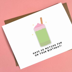 May include: A white greeting card with a matcha drink illustration. The drink is green with a pink topping and a brown stick. The card reads "HAVE SO MATCHA FUN ON YOUR BIRTHDAY!" on a pink background.