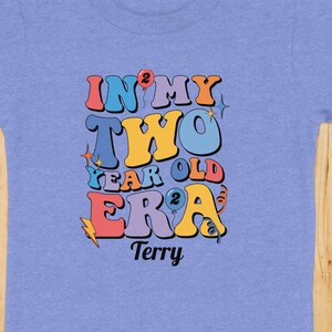 May include: A light blue toddler t-shirt with the text "IN MY TWO YEAR OLD ERA" in colorful retro font. The name "Terry" is printed below the text. The shirt is on a wooden surface with a hat, jeans, a purse, and a toy airplane.