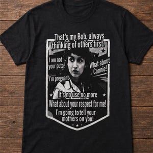 May include: Black t-shirt featuring a black and white image of a woman with text that reads "That's my Bob, always thinking of others first." Additional text includes phrases like "I am not your puta!" and "It's no use no more."