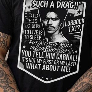 May include: Black t-shirt with a graphic design featuring a portrait and text. The text includes "STOP BEING SUCH A DRAG!!" and "LUBBOCK TX!?", along with other phrases. The shirt is worn by a person with tattooed arms.