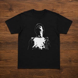 May include: Black t-shirt featuring a white graphic of a person looking upwards. The graphic is a stylized, minimalist design with a focus on the silhouette and the upward gaze. The shirt is made of a soft material.